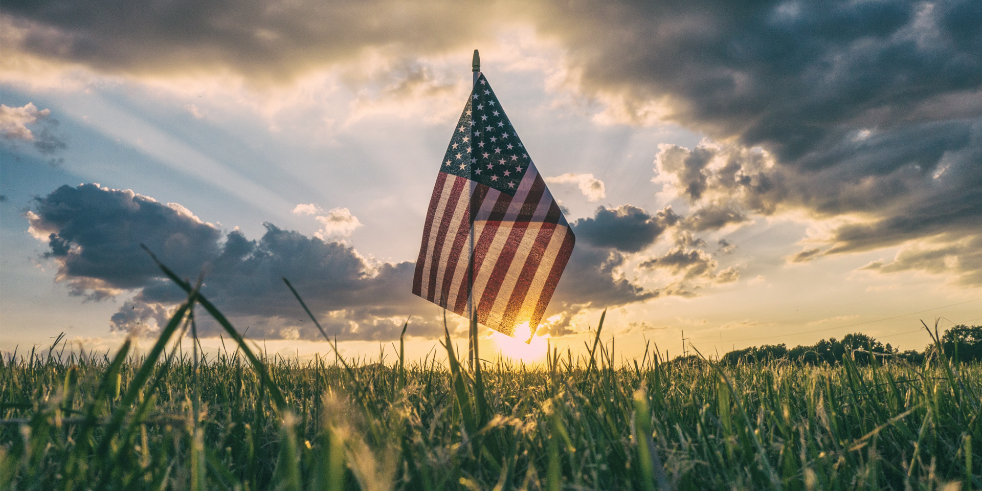 American flag in grassy ground with sun and clouds in sky